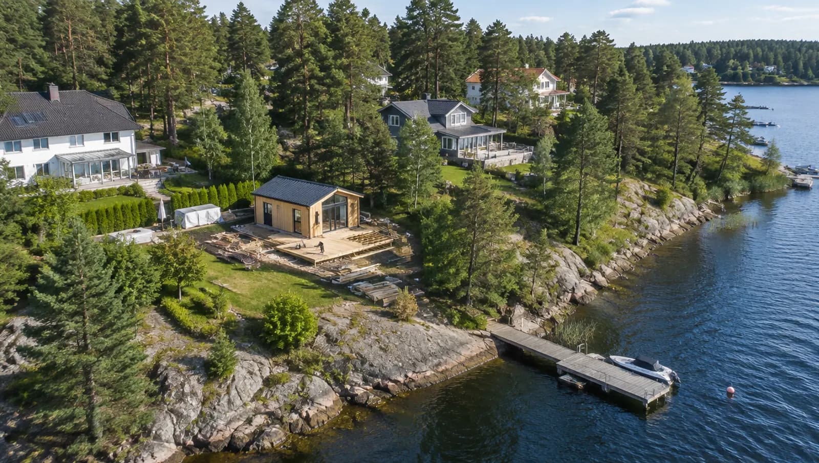 Flygbild över ett villaområde bland tallar vid en sjövik utanför Stockholm, där ett litet attefallshus byggs i en trädgård